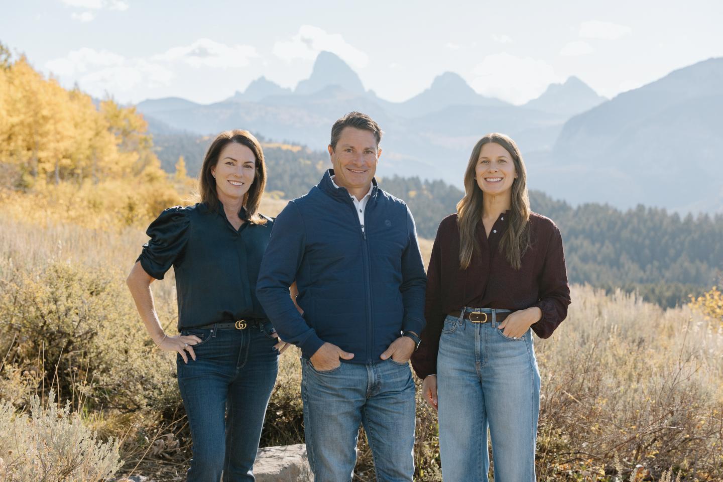 Three people smiling, standing in a scenic mountainous landscape with autumn foliage.